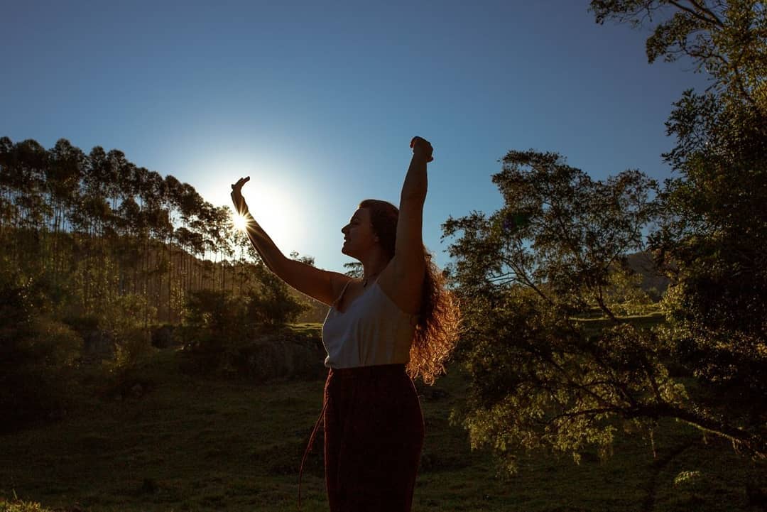 Aline Ferreira Gomes em campo aberto ao entardecer, braços erguidos contra o sol entre árvores