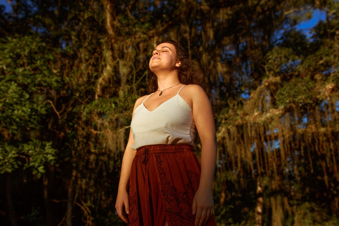 Aline em pé entre árvores ao entardecer dourado, cabeça levantada e olhos fechados em respiro profundo