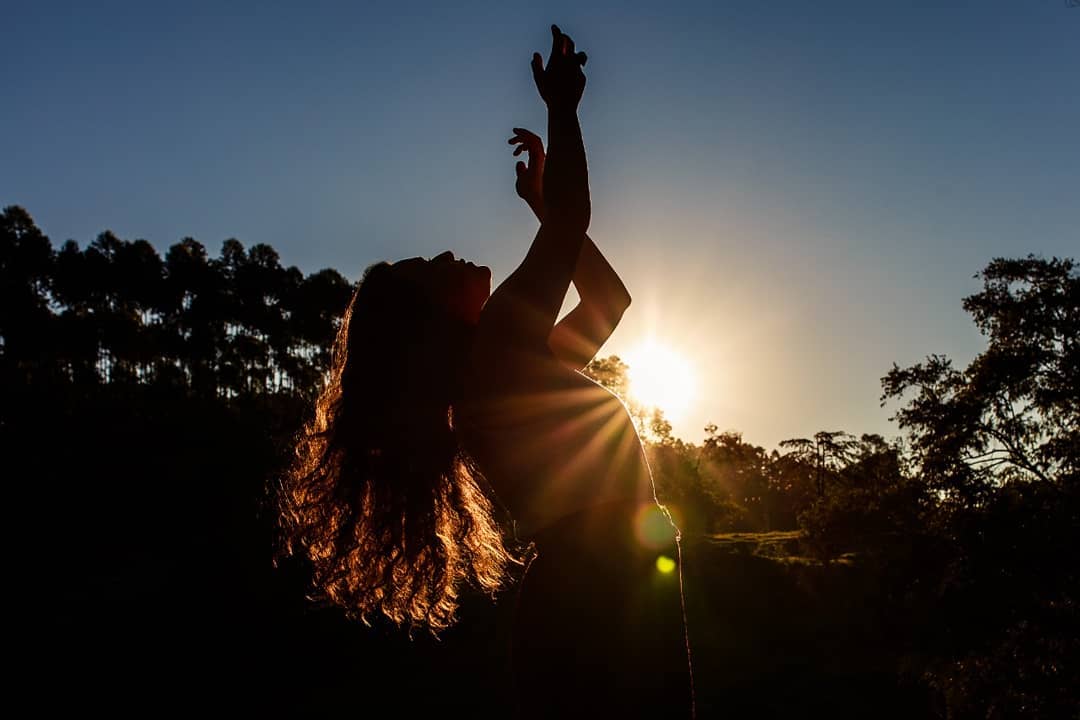 Silhueta de Aline em contraluz contra um pôr-do-sol intenso, braços erguidos em gesto de potência