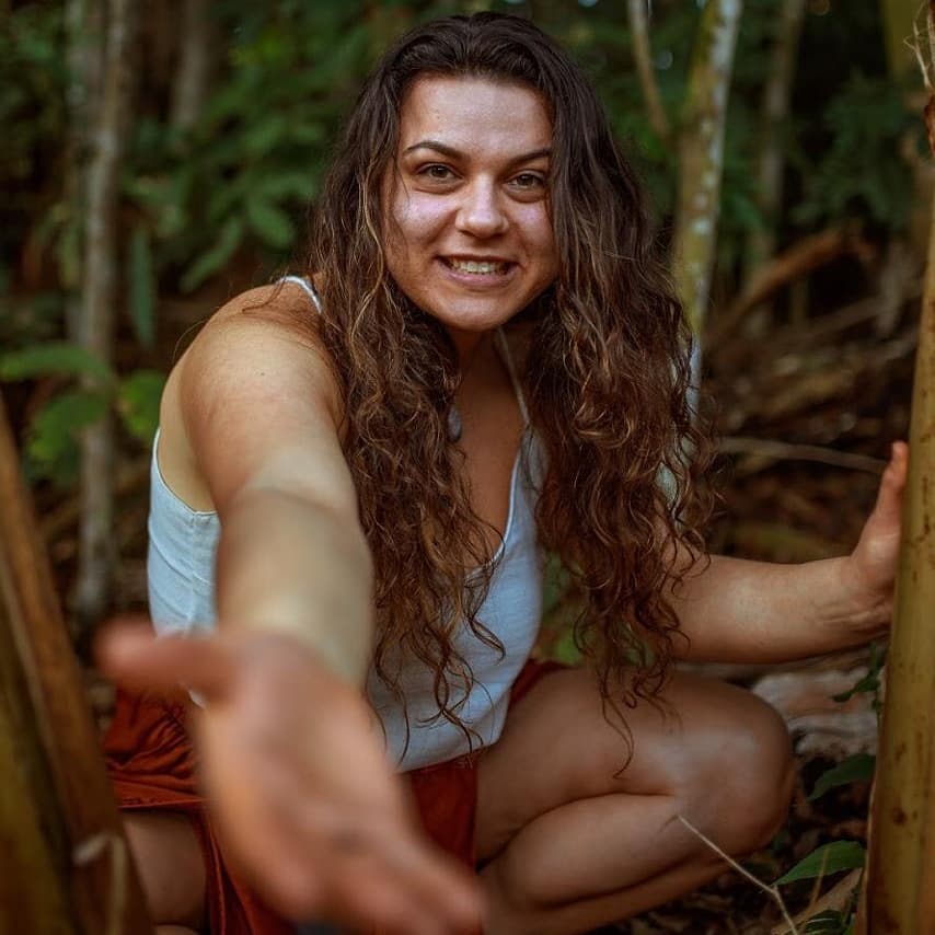 Aline agachada entre bambus na mata, sorrindo e estendendo o braço pra frente em gesto de oferta
