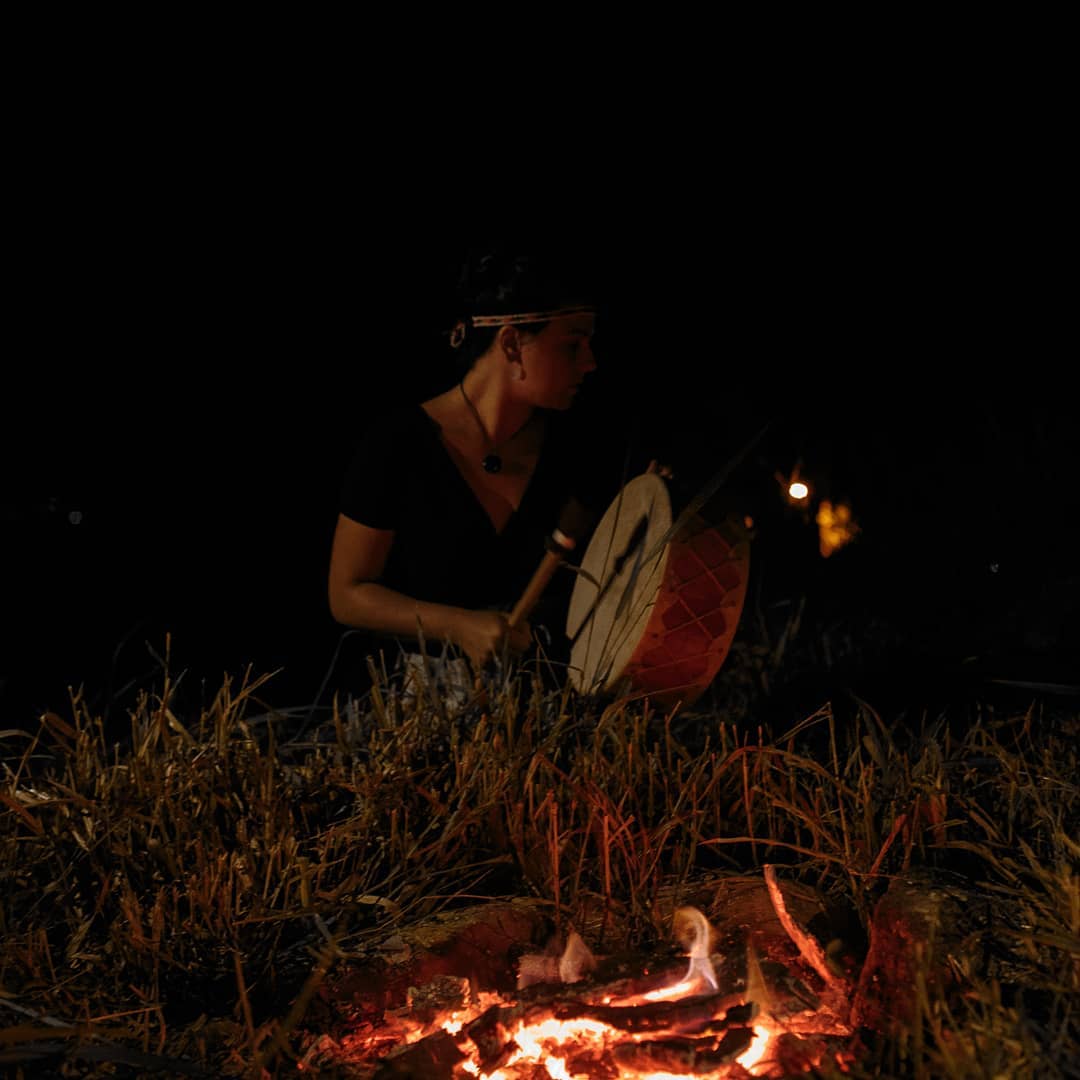 Aline tocando um tambor xamânico à noite, próxima a uma fogueira em sessão de cura