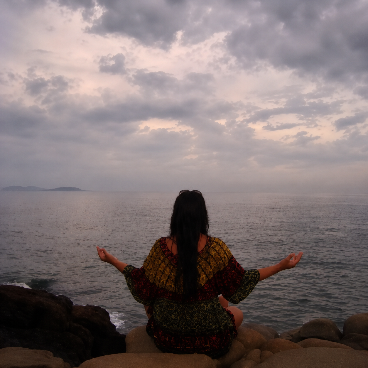 Mulher de costas em postura de meditação sentada em pedras à beira-mar, olhando o horizonte de céu nublado